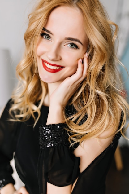 Closeup portrait of smiling young woman with long blonde wavy hair. Wearing elegant black blouse, dress with sequins, light day makeup with red lipstick.