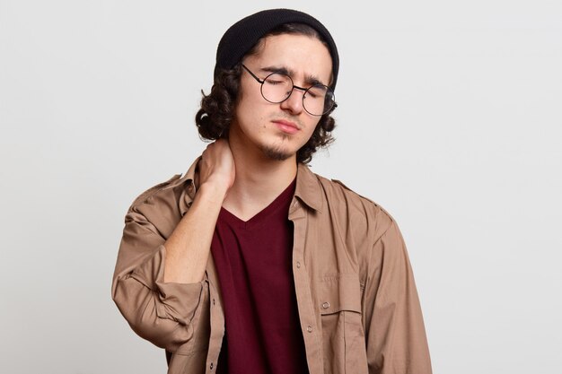 Closeup portrait of stressed exhausted youngster touching her neck with hand, doing massage, having neck pain, looks tired, closing his eyes, eager to have rest, wearing casual clothes. Youth concept.