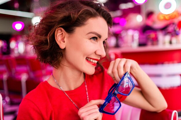 Крупным планом портрет стильной улыбающейся женщины в красочном наряде в ретро-винтажном кафе 50-х годов, сидящей за столом в красной рубашке, синих солнцезащитных очках, веселящейся в веселом настроении с красной помадой