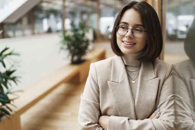 Foto gratuita ritratto del primo piano di una giovane donna di successo e sicura di sé che inizia la carriera con un aspetto determinato a ottenere un colloquio in attesa di lavoro appoggiato sulla parete di vetro nel centro business sorridente e distogliendo lo sguardo soddisfatto