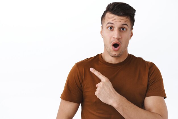Closeup portrait of surprised and excited shocked handsome man open mouth pointing finger left at something intriguing and unique promoting new product in stock white background