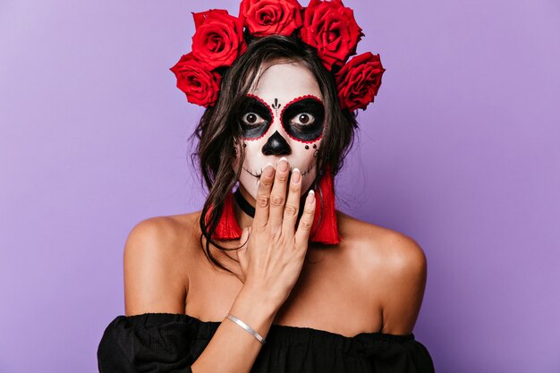 Портрет крупного плана удивленной девушки с карими глазами и косметикой для Хэллоуина. Взрослая женщина с короной из роз прикрывает рот рукой от шока.