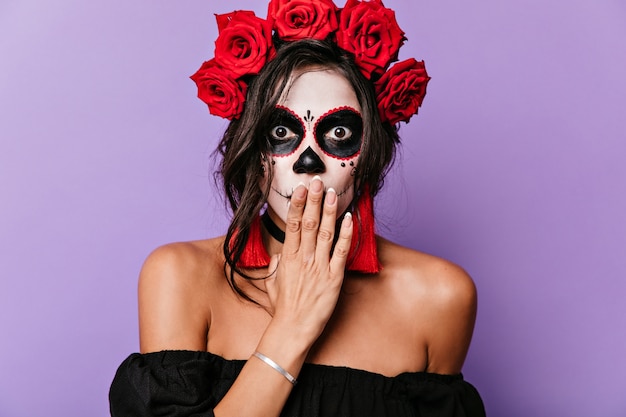 Closeup portrait of surprised girl with brown eyes and makeup for Halloween. Adult woman with crown of roses covers her mouth with her hand from shock.