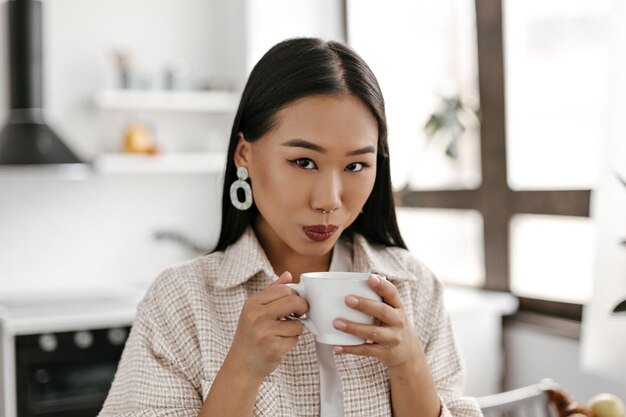 日焼けしたブルネットのアジアの女性のクローズアップの肖像画と暗い口紅はお茶を飲みます女性はカメラをのぞき、キッチンで白いコーヒーカップを保持します