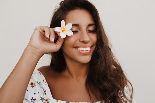 Closeup portrait of tanned lady with snow-white smile posing with flower on white wall