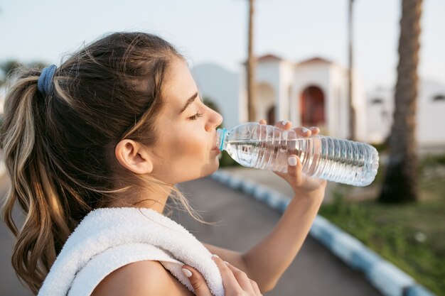 晴れた朝の通りのトレーニング中にボトルから水を飲んでのどが渇いて若いきれいな女性のクローズアップの肖像画。陽気な気分、目を閉じて身も凍るよう、夏、フィットネスタイム