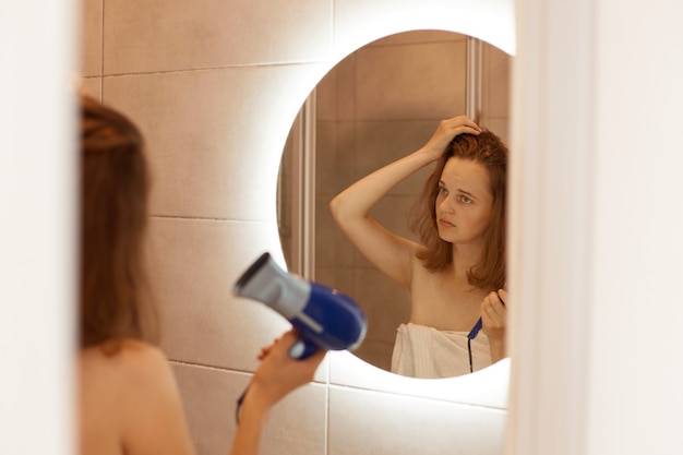 Free photo closeup portrait of an unhappy young woman looking at her damaged hair, holding hair dryer, looking at her reflection in mirror with sad facial expression.