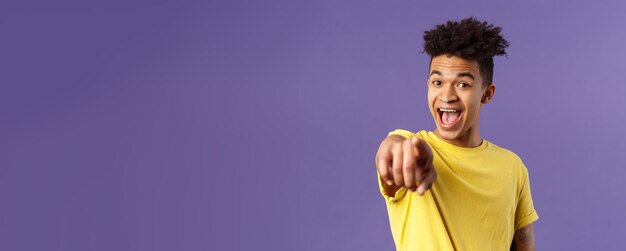 Ritratto del primo piano dell'uomo ispanico stupito ottimista con il dito puntato del giovane studente di dreadlocks alla macchina fotografica a