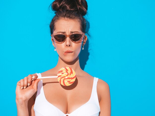 Closeup portrait of young beautiful sexy woman with ghoul hairstyle. Trendy girl in casual summer white swimsuit in sunglasses.Hot model isolated on blue.Eating,biting candy lollipop