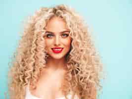 Free photo closeup portrait of young beautiful smiling blond hipster female in trendy summer clothes sexy carefree woman posing near blue wall with afro curls hairstyle positive model having fun