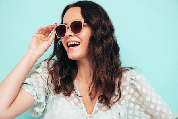 Closeup portrait of young beautiful smiling female in trendy summer dress Sexy carefree woman posing near blue wall in studio Positive model having fun indoors Cheerful and happy In sunglasses