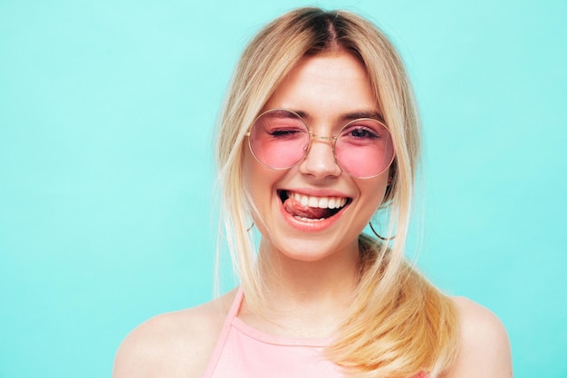 Free photo closeup portrait of young beautiful smiling female in trendy summer hipster clothes sexy carefree woman posing on blue background in studio positive model winks and shows tongue in sunglasses