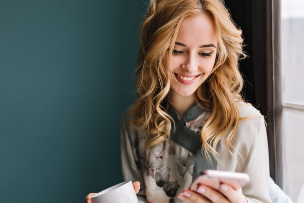 コーヒーやお茶を飲みながら電話でウェーブのかかった髪のテキストメッセージを持つ若いブロンドの女性のポートレート、クローズアップ。彼女はターコイズブルーの壁の明るい部屋にいます。レースパジャマを着用。