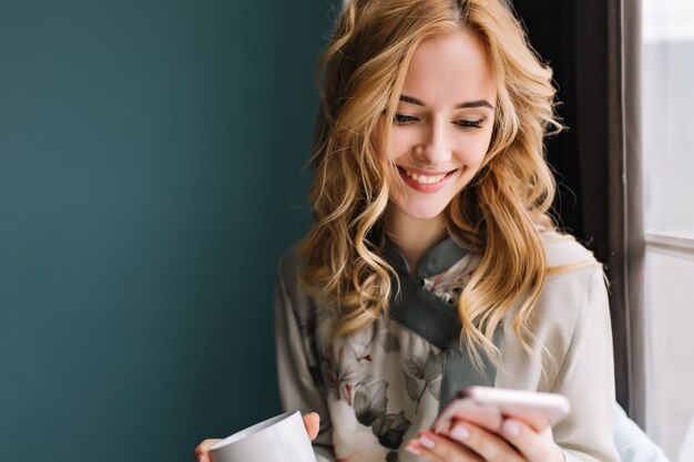 Макрофотография портрет молодой блондинки с волнистыми волосами текстовых сообщений на телефоне, пить кофе или чай. Она находится в светлой комнате с бирюзовой стеной. В кружевной пижаме.