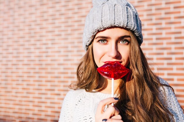 Free photo closeup portrait young girl with long hair in knitted hat with caramel red lips on wall  outside. she is looking .