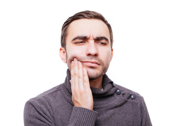 Closeup portrait of young man with tooth ache crown problem about to cry from pain touching outside mouth with hand, isolated on white wall