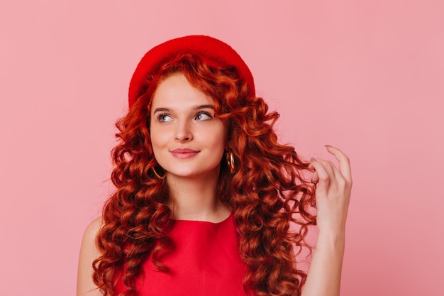 Closeup portrait of young stylish woman in red beret and top Blueeyed girl dreamily touches her red curls on pink background Closeup portrait of young stylish woman in red beret and top