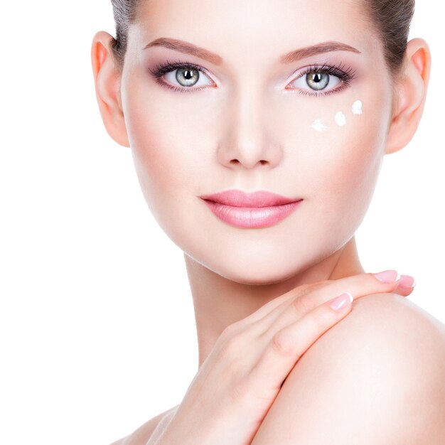 Closeup portrait of young woman applying cream on her pretty face - on a white wall.