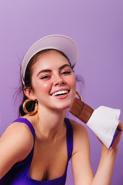 明るい金のイヤリングと紫色の壁にチョコレートのバーを笑顔と保持している白い帽子の若い女性のクローズアップの肖像画