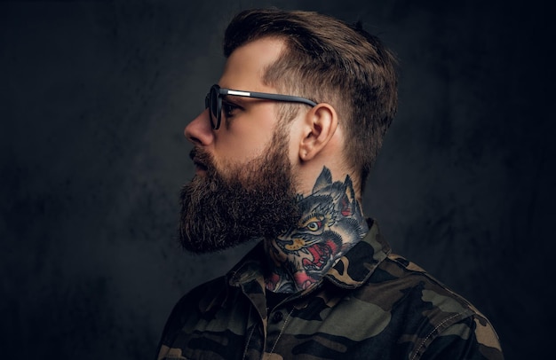 Free photo closeup profile of a bearded man with a tattoo on his neck in sunglasses wearing a military shirt. studio photo against a dark wall