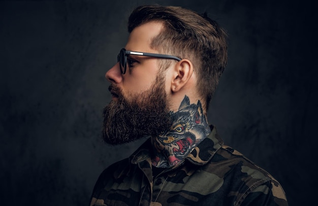 Closeup profile of a bearded man with a tattoo on his neck in sunglasses wearing a military shirt. Studio photo against a dark wall