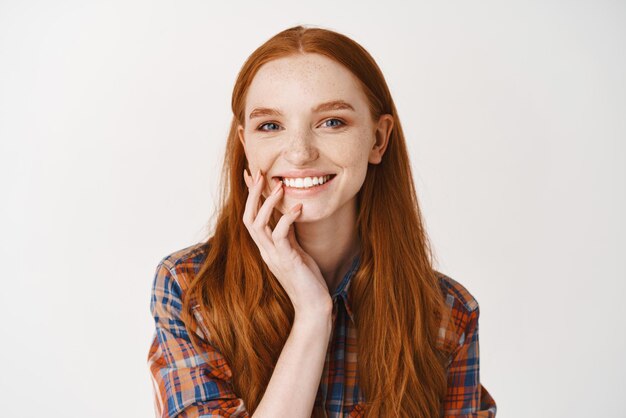 Closeup of redhead female without makeup and white perfect smile looking happy at camera standing against white background