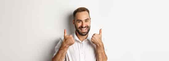 Free photo closeup of satisfied bearded guy in white shirt showing thumbs up in approval like and agree positiv