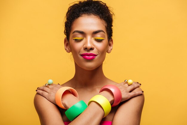Closeup satisfied naked mulatto woman with fashion makeup and accessories posing on camera with crossed hands on shoulders, over yellow