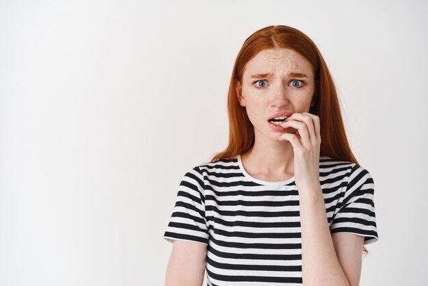 Closeup of scared and insecure redhead girl biting fingernails looking worried at camera standing over white background