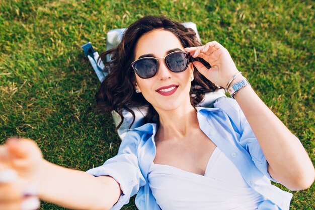 Селфи-портрет крупного плана милой девушки брюнет с короткими волосами кладя на траву в парке. Она носит белую футболку и синюю рубашку, солнцезащитные очки. Вид сверху.