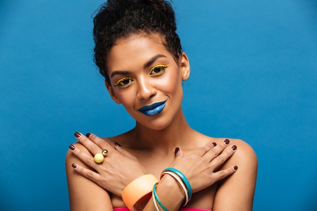 Closeup sensual naked mulatto woman with fashion makeup and accessories with crossed hands on chest, over blue wall