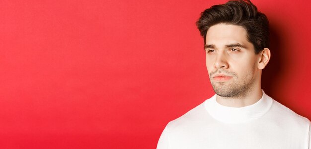 Closeup of serious and thoughtful man in white sweater looking left standing against red background