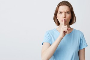 Closeup of serious unhappy young woman in blue t shirt frowning face and showing silence sign isolated on white