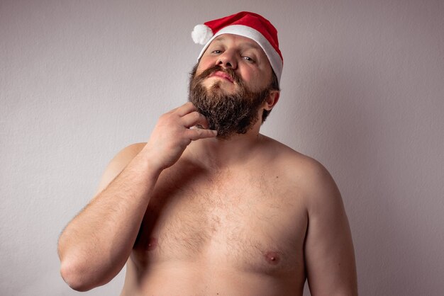 Крупным планом бородатый мужчина без рубашки в шляпе Санта-Клауса на сером фоне