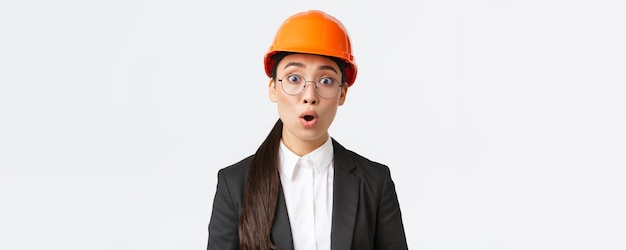 충격을 받고 걱정스러운 아시아 여성 엔지니어의 클로즈업은 안전 마스크 안경과 비즈니스 정장에 서서 놀란 흰색 배경에 서서 걱정스러운 발표를 듣는 나쁜 소식을 듣습니다.