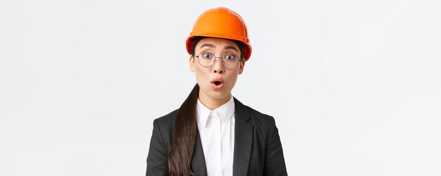 ショックを受けて心配しているアジアの女性エンジニアのクローズアップは、安全マスクの眼鏡とビジネススーツに立って、びっくりした白い背景をあえぎながら、悪いニュースを心配するアナウンスを聞きます