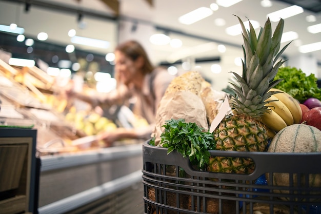 Крупным планом тележка для покупок в супермаркете, полном продуктов, фруктов и овощей, в то время как на заднем плане женщина снимает продукт с полок