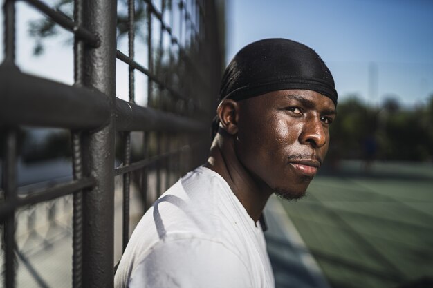 Closeup shot of an African-American male in a white shirt leaning on a fence at the basketball court