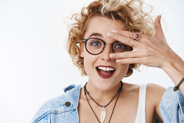Closeup shot of charismatic happy and enthusiastic stylish young emotive woman with blond short haircut in glasses and denim jacket open mouth joyful and carefree looking through fingers surprised
