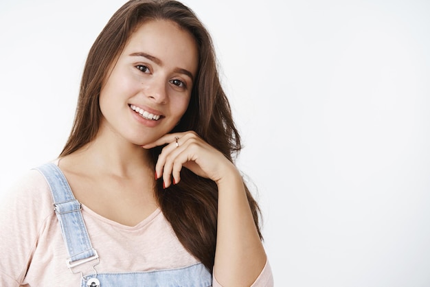 Closeup shot of charming tender and cute female brunette in casual outfit gently touching jawline and smiling broadly gazing dreamy at camera liking new facial mask that making skin pure