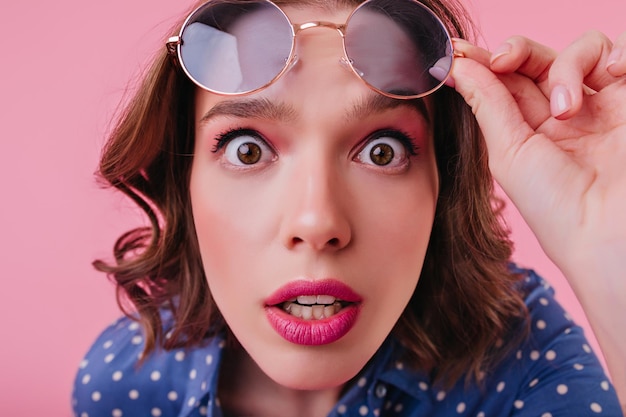 Free photo closeup shot of darkhaired surprised female model holding her sunglasses portrait of amazed white girl isolated on pink background with mouth open