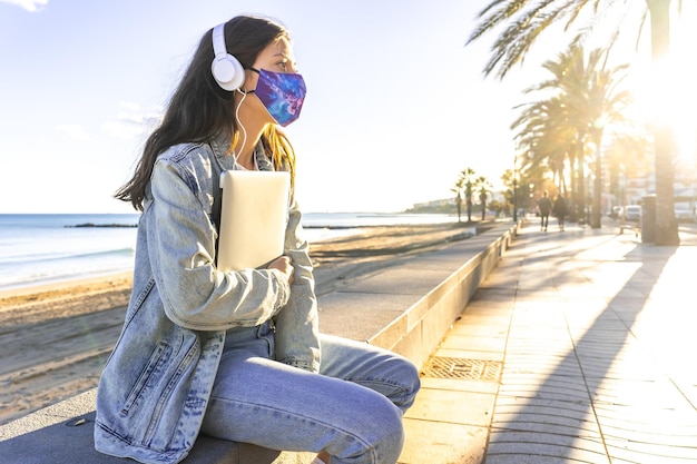 Closeup shot of a female in a mask, headphones and laptop in hands sitting on the curbstone