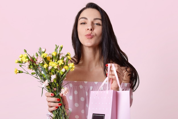 Closeup shot of good looking dark haired young woman keeps lips folded, holds gift bag and flowers, gives present to friend