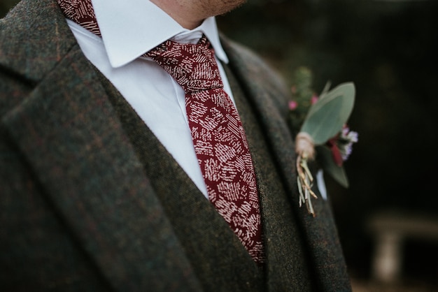 Free photo closeup shot of a groom's suit with flowers and red patterned tie with trees on the background