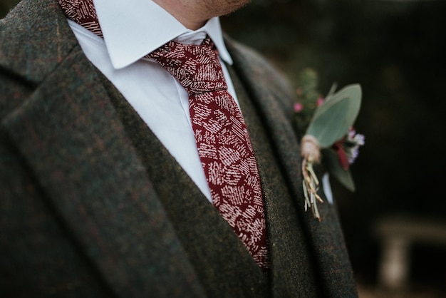 Closeup shot of a groom's suit with flowers and red patterned tie with trees on the background