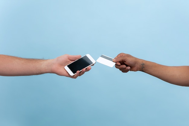 Closeup shot of human holding hands isolated on blue wall. Relations, friendship, partnership, business or family. Copyspace. Holding a card and a smartphone. Online payments bill.