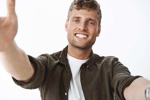 Colpo del primo piano di amorevole e premuroso ragazzo bello tenero con gli occhi azzurri e ampio sorriso bianco tirando le braccia verso la telecamera per coccolarsi o abbracciarsi che vogliono tenersi in mano in posa felice sul muro grigio