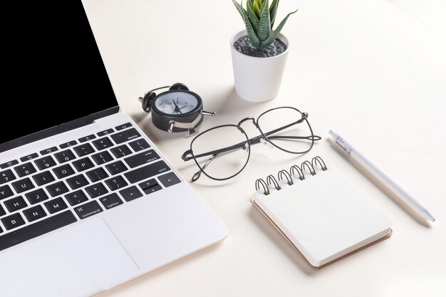 Closeup shot of an open laptop, glasses, a plant, a pen, a notepad, and an alarm clock