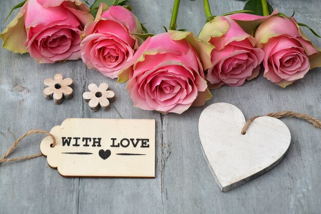 Closeup shot of pink roses next to an empty heart wooden tag and a with love tag on a wooden surface