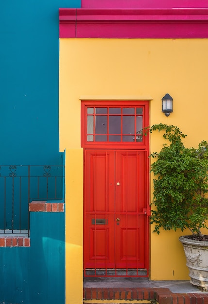 Closeup shot of the red door of a yellow building and a plant next to it